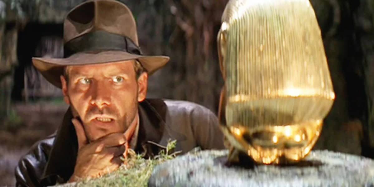 A person in a brown hat and leather jacket closely observes a golden idol on a stone pedestal in a dimly lit setting.