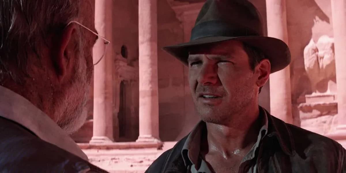 A man wearing a brown fedora and leather jacket stands facing another man with gray hair and glasses. They appear to be having a serious conversation in front of a large ancient stone structure with columns.