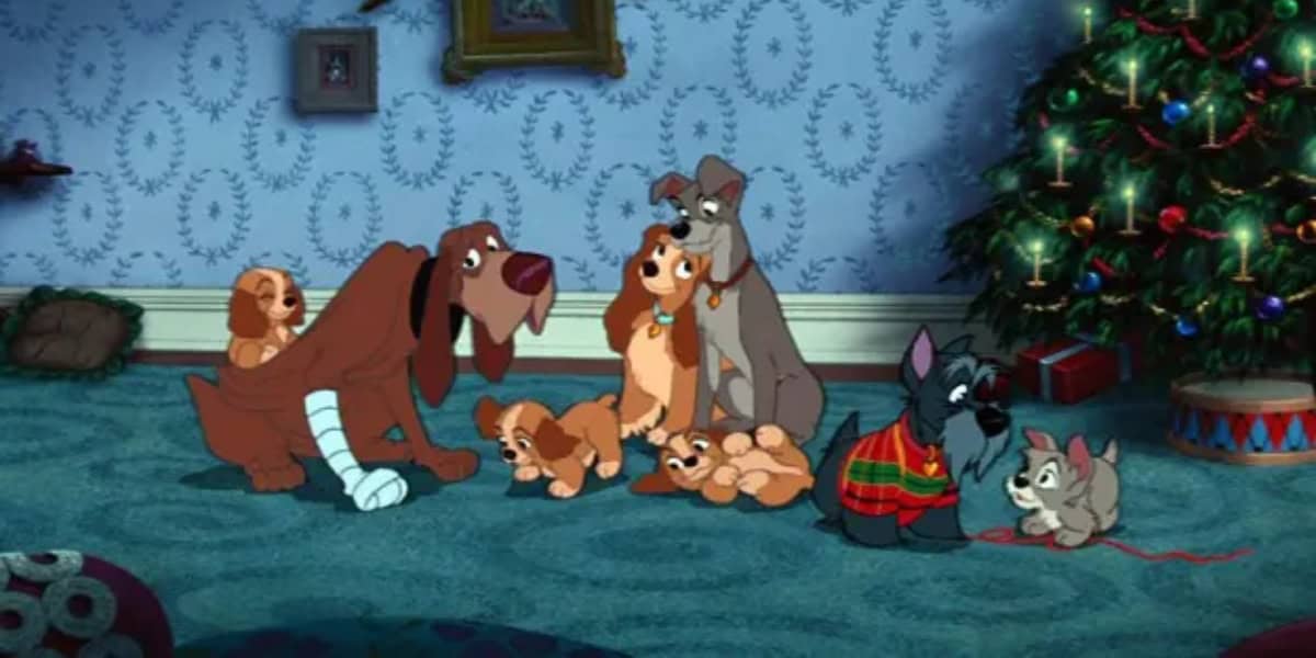 An animated scene featuring several dogs sitting on a carpeted floor next to a decorated Christmas tree. A dog with a bandaged paw sits near puppies, while another larger dog stands beside a smaller one wearing a red sweater, playing with a cat.