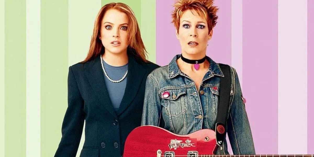 Two women stand side by side against a striped background. The one on the left wears a suit and pearls, while the one on the right sports a denim jacket and holds a red electric guitar. Their expressions appear surprised.
