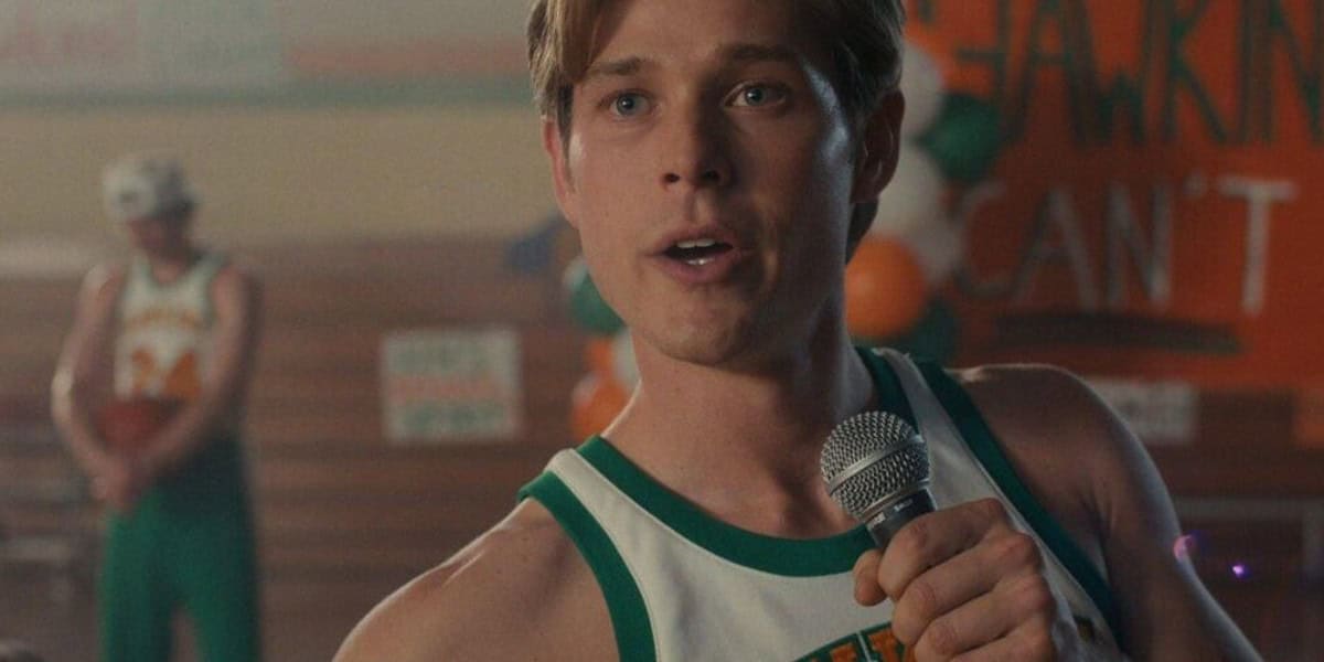 A young man in a basketball jersey holds a microphone, speaking at an indoor event. Another person stands blurred in the background. The venue is decorated with colorful balloons and a partially visible banner.