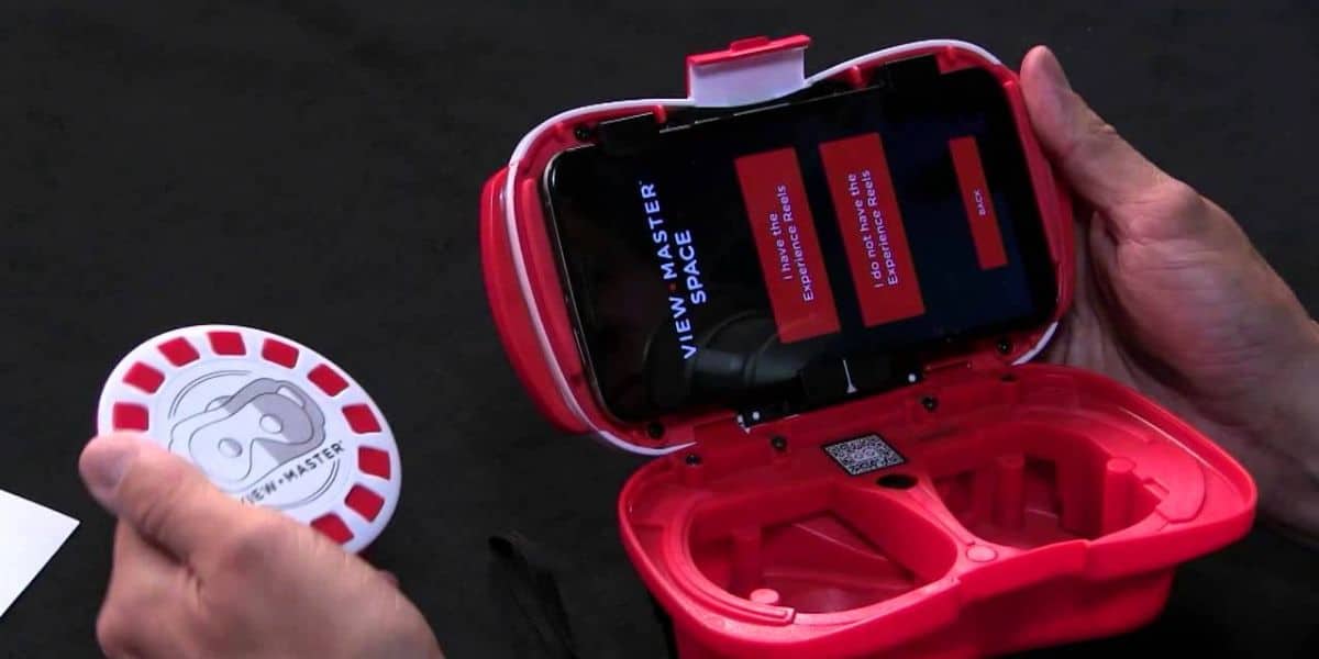 A person holds a red virtual reality viewer, open to show a smartphone screen inside with the text "VIEW-MASTER SPACE." Another hand holds a round card with images, resembling a classic View-Master reel.