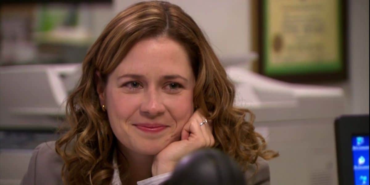 A person with curly hair is smiling softly, resting their chin on their hand. They are seated at a desk with office equipment and a framed certificate in the background.