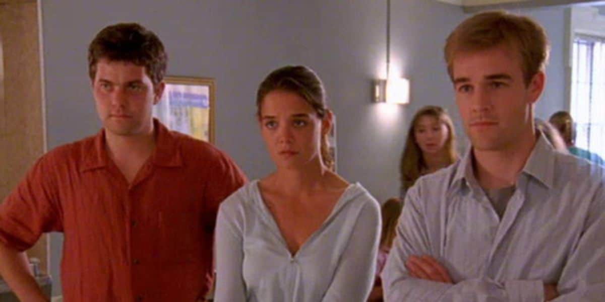 Three people stand indoors, two men and a woman, all looking serious. The man on the left wears a red shirt, the woman wears a light blue top, and the man on the right wears a light gray shirt. Another person is blurred in the background.
