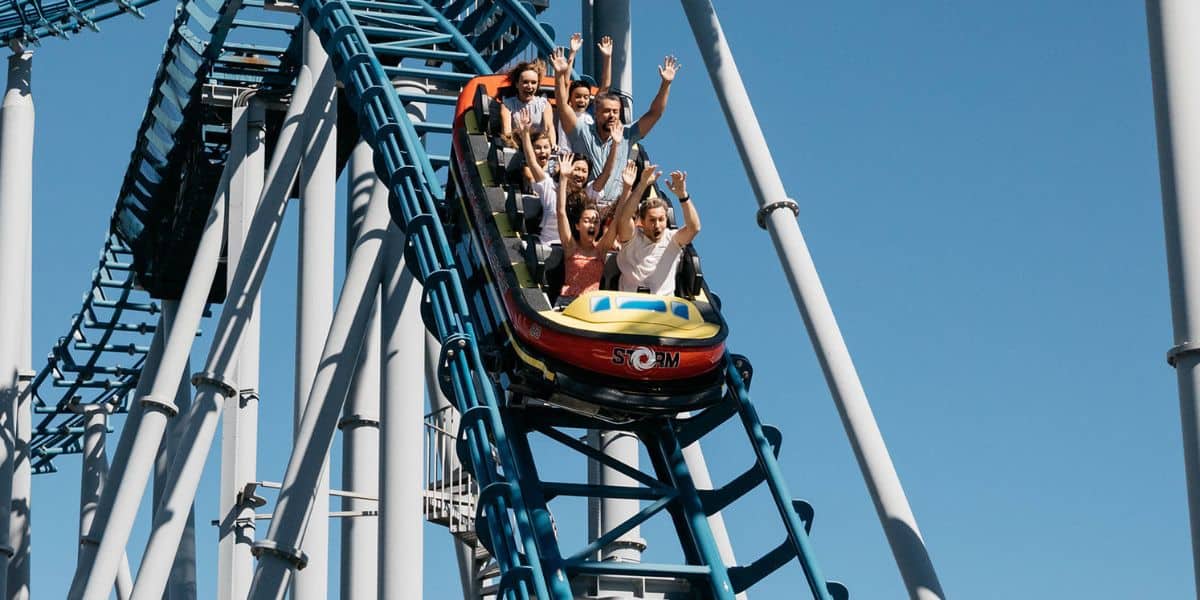 A group of people enjoy a ride on a roller coaster, raising their hands as they descend a steep track. The sky is clear and blue, and the roller coaster is painted with bright colors.
