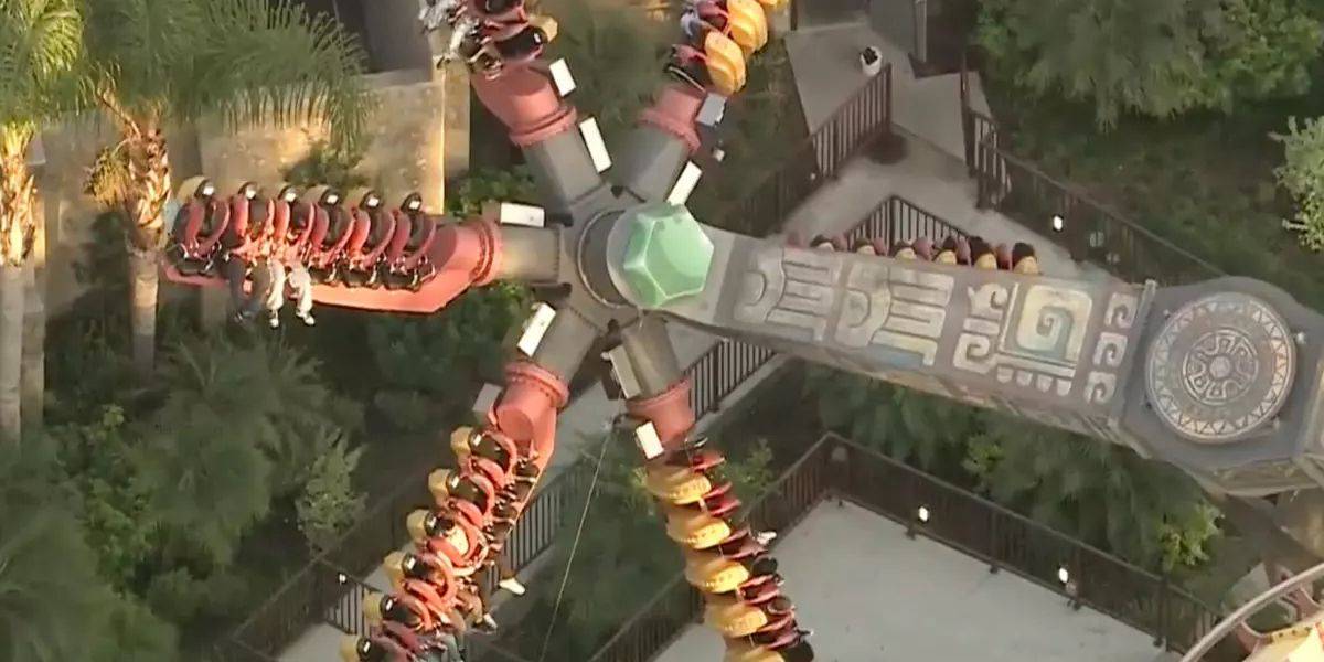 Aerial view of people on a large spinning amusement park ride with four arms, each holding a row of seated riders. The ride features decorative patterns and is set amidst greenery on a sunny day.