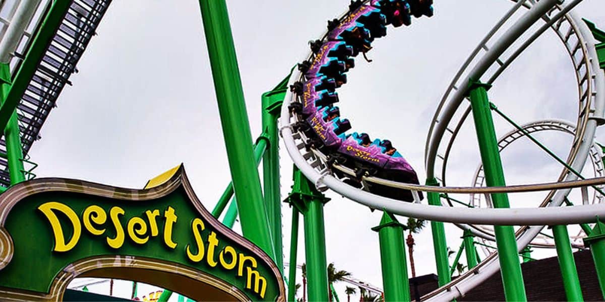 A vibrant roller coaster named Desert Storm with green tracks and bright purple cars, loops in the air against a cloudy sky. The neon sign at the entrance stands out in the foreground. Palm trees are visible in the background.