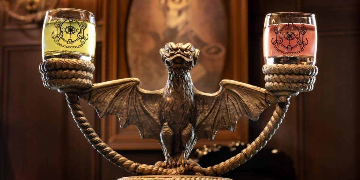 A stone gargoyle sculpture holds two glasses filled with yellow and pink liquids, each emblazoned with a dragon emblem. The scene is set against a rich wooden backdrop with a framed portrait on the wall.