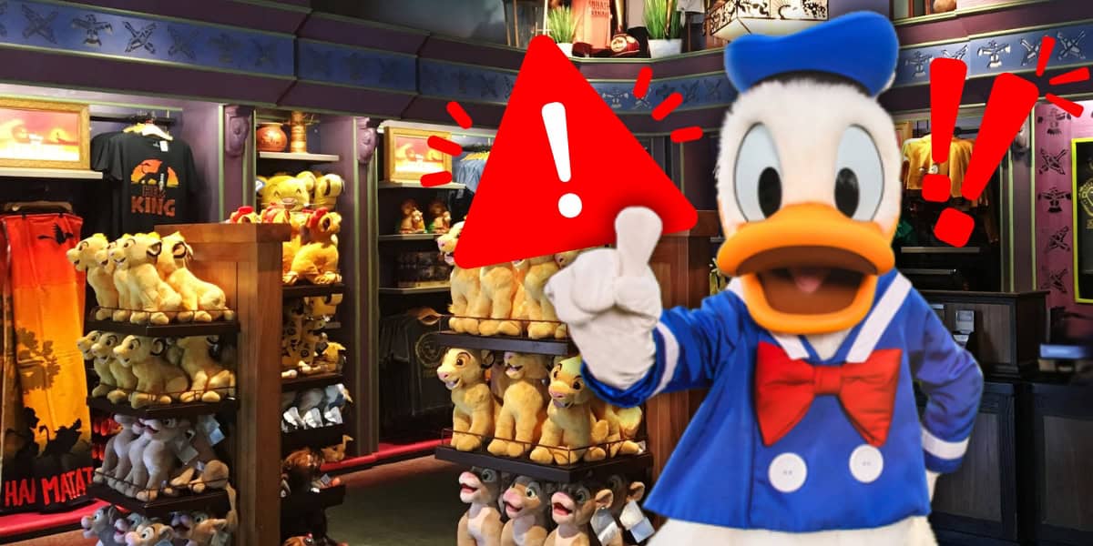 A person in a Donald Duck costume stands in a store filled with plush toys and Disney merchandise. They point to a large red alert sign with an exclamation mark. Merchandise includes stuffed animals and colorful clothing.