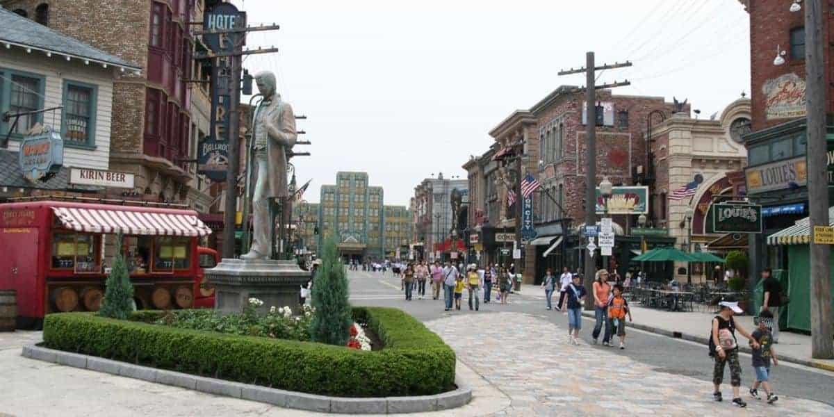 A lively street scene at Universal Studios Japam s featuring vintage-style buildings, a statue on a landscaped median, and various flags and signage. Visitors stroll along the cobblestone path, enjoying the attractions and food stands.