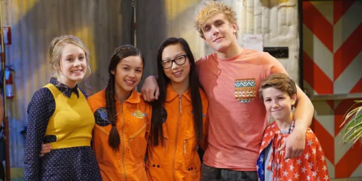 A group of five young people stand together smiling. Two wear orange uniforms, two casual clothes, and one in a polka dot dress with a yellow top. They pose in a colorful, indoor setting.