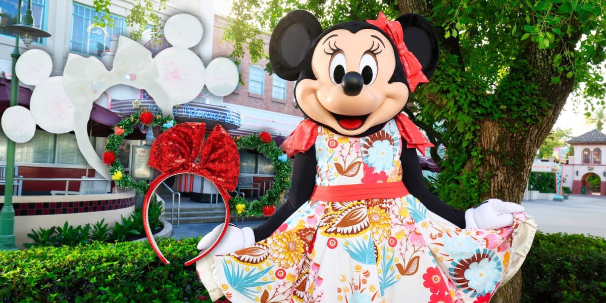 Minnie Mouse, dressed in a colorful floral dress and red headband with a bow, stands happily in a theme park setting. There's a large bow sculpture adorned with flowers in the background, and trees are casting some shade.