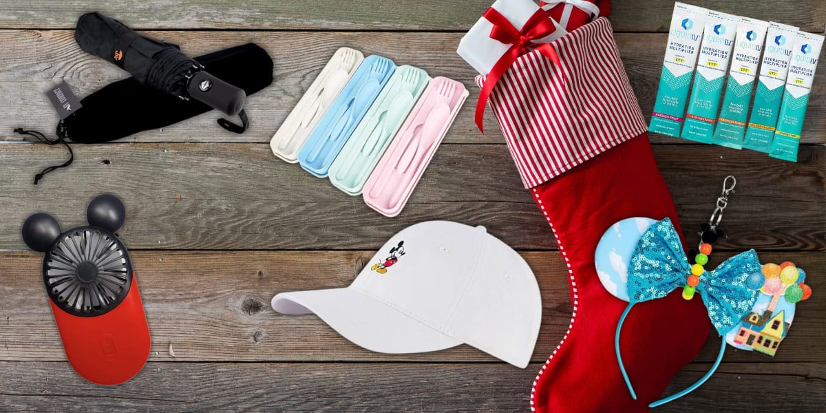 A festive stocking filled with various items on a wooden surface. Contents include a white cap with a small design, pastel-colored stacked socks, an umbrella, a portable fan, bright blue mouse ears, and several packs of allergy medicine.
