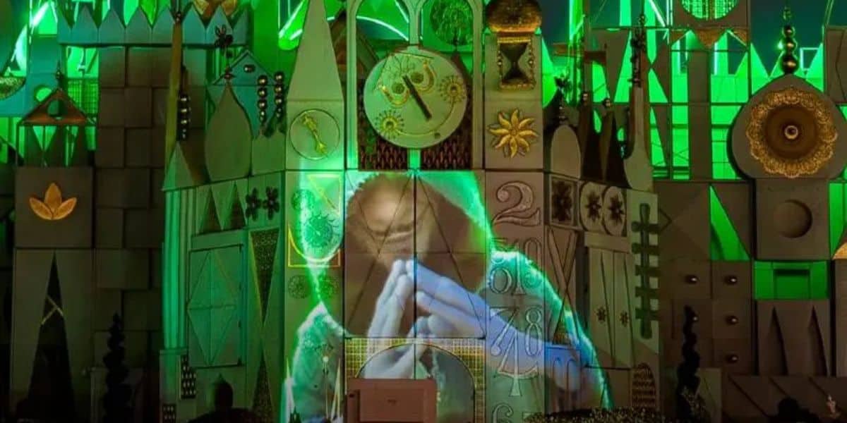 A vibrant green-lit facade with various shapes and designs, featuring a projected image of a person with clasped hands, possibly in prayer or meditation. The scene includes a variety of geometric patterns and clock-like elements.