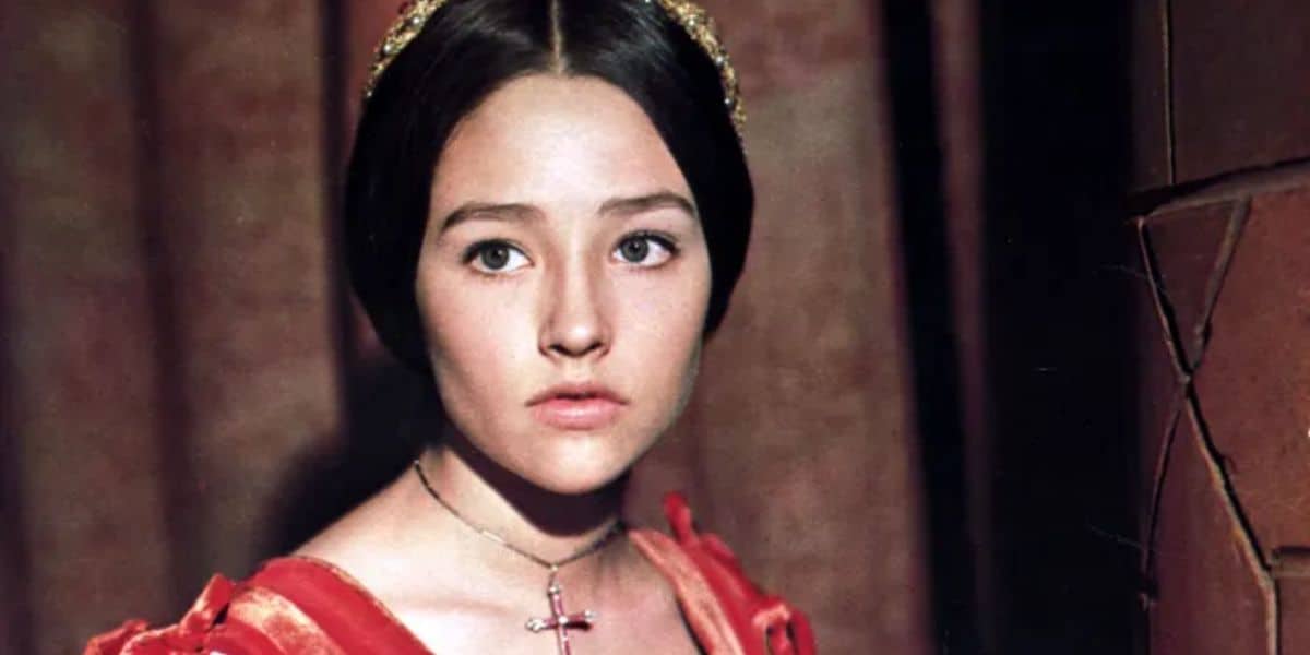 A young woman with dark hair looks slightly to the side. She wears a red dress with ruffled sleeves and a cross necklace. Her expression is thoughtful, and a stone wall is in the background.