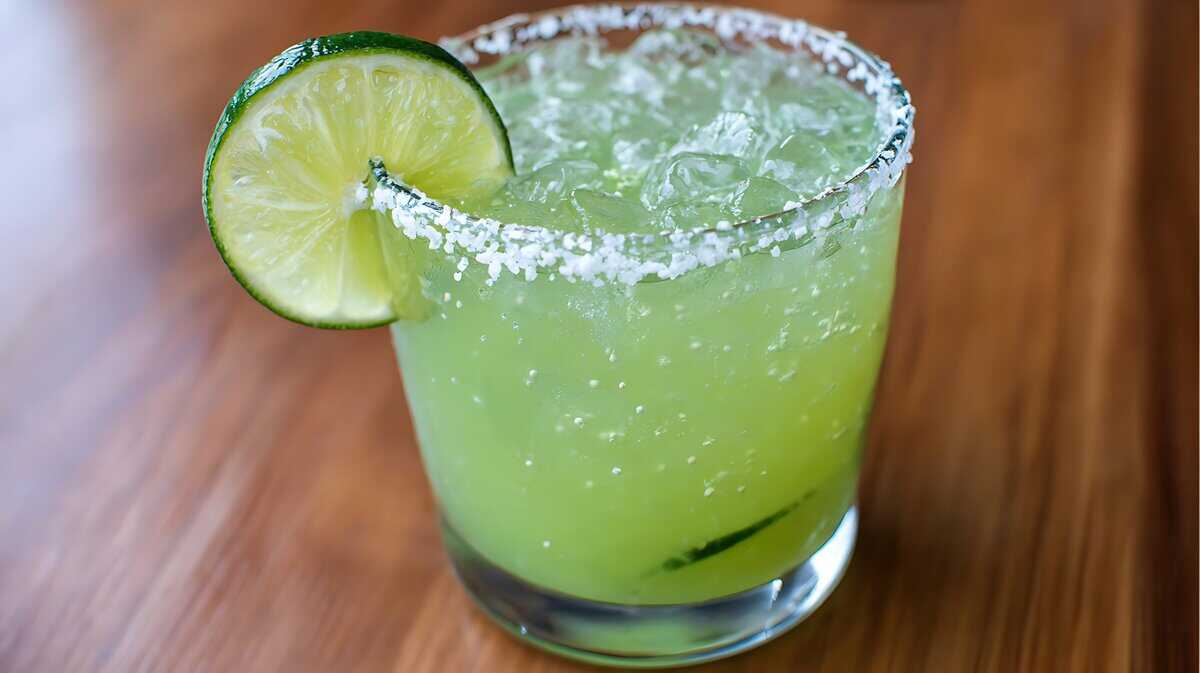 A glass of margarita inspired by La Cava del Tequila recipes, with a salted rim, filled with ice and garnished with a fresh lime slice, sits on a wooden surface.