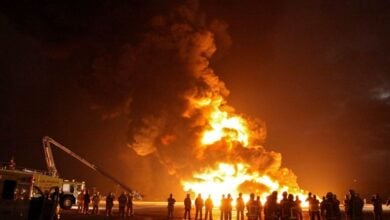 Silhouetted firefighters stand in front of a massive blaze, with large flames and thick smoke billowing into the night sky. A fire truck with an extended hose ladder is visible on the left.