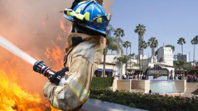 A firefighter wearing protective gear sprays water on a blaze with flames in the background. In the distance, there is an entrance with a sign saying "Universal Studios" surrounded by palm trees.