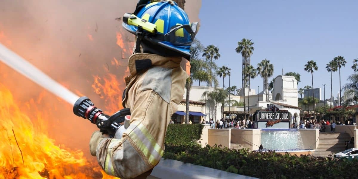 A firefighter wearing protective gear sprays water on a blaze with flames in the background. In the distance, there is an entrance with a sign saying "Universal Studios" surrounded by palm trees.