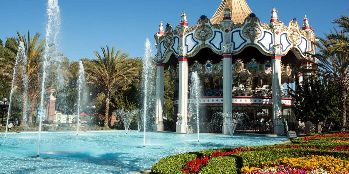 A vibrant amusement park scene featuring a large, ornate carousel with decorative patterns. In the foreground, water fountains spray into a clear, blue pool surrounded by palm trees and lush flowerbeds under a sunny sky.