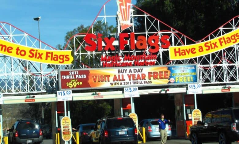 Entrance to Six Flags Magic Mountain with a colorful sign, featuring a roller coaster backdrop. Banners indicate ticket prices and promotions, including a 2011 thrill pass offer. Cars are lined up at the booths beneath the vibrant signage. Six Flags Magic Mountain reopens