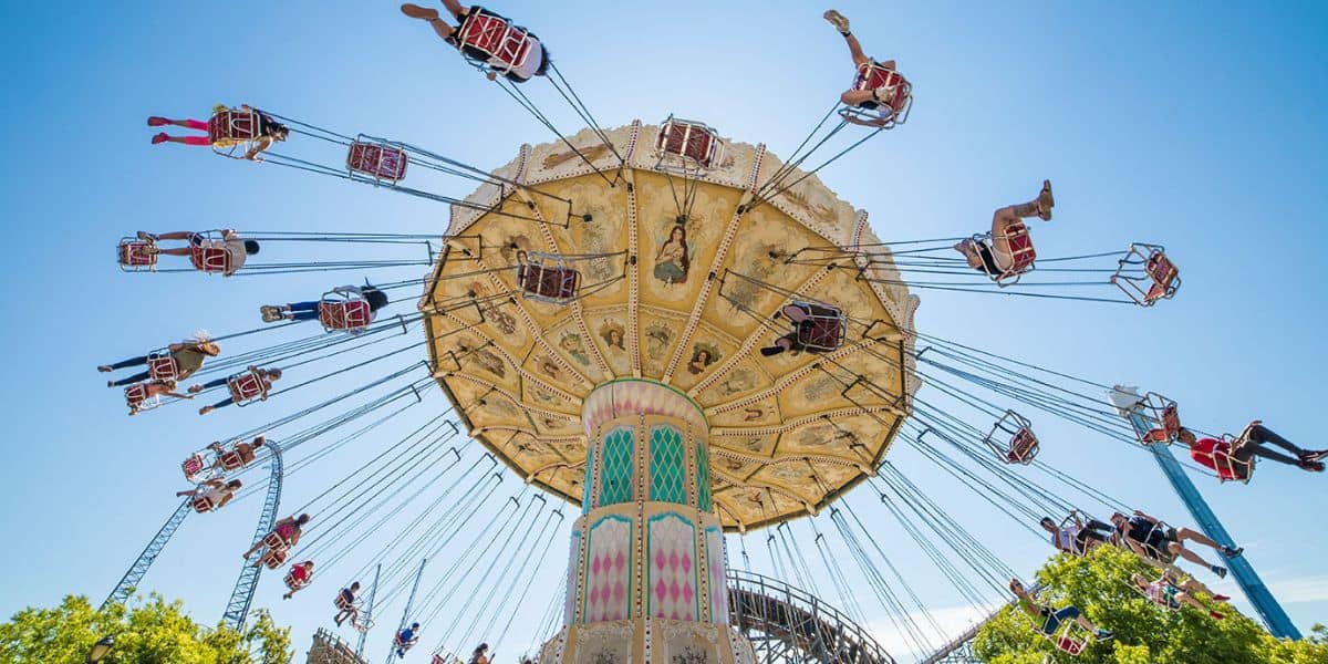 A brightly colored swing ride at an amusement park spins with people sitting in seats suspended by chains, flying through the air against a clear blue sky. Trees and roller coaster tracks are visible in the background.