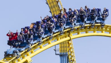 People ride a roller coaster with blue seats and yellow tracks, experiencing excitement and thrill. Some riders have their arms raised, while others hold onto the rail. The sky is clear, emphasizing the height of the loop, where theme park news means no more summer tickets for 2025.