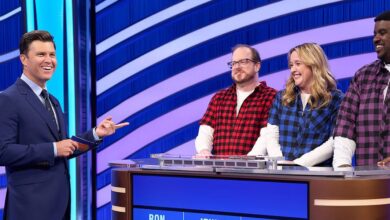 A game show host in a navy suit interacts with three contestants standing behind a podium. The contestants wear plaid shirts in red, blue, and purple. A digital display shows names and scores. The background features blue and white curved stripes.