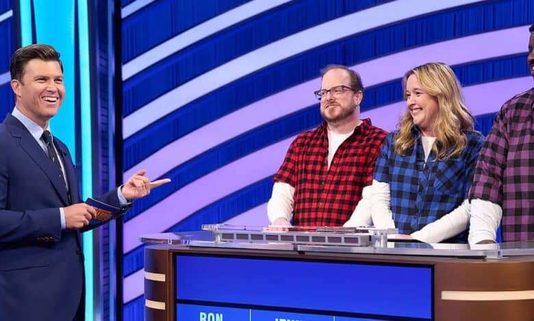 A game show host in a navy suit interacts with three contestants standing behind a podium. The contestants wear plaid shirts in red, blue, and purple. A digital display shows names and scores. The background features blue and white curved stripes.