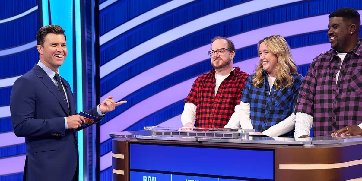 A game show host in a navy suit interacts with three contestants standing behind a podium. The contestants wear plaid shirts in red, blue, and purple. A digital display shows names and scores. The background features blue and white curved stripes.