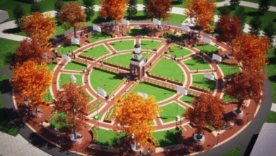 Aerial view of a circular park with a central clock tower, surrounded by pathways. Autumn-colored trees are evenly spaced, and people are walking or sitting on benches. The grassy areas and pathways form a symmetrical pattern.