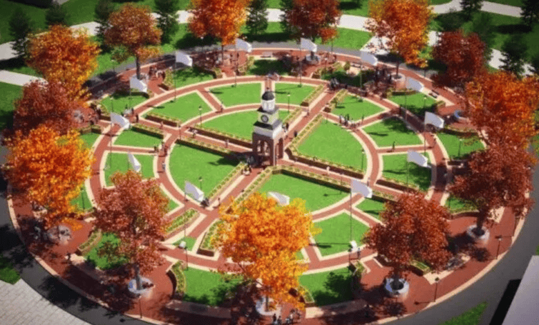Aerial view of a circular park with a central clock tower, surrounded by pathways. Autumn-colored trees are evenly spaced, and people are walking or sitting on benches. The grassy areas and pathways form a symmetrical pattern.