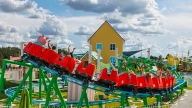 A bright red roller coaster with excited riders passes a yellow house at an amusement park. The track is vibrant green, and the sky is partly cloudy. In the background, there are trees and other colorful structures.