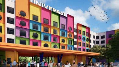 A colorful building with a large "Universal" sign features a facade of bright squares in various colors. Circular windows are set within the squares. People walk in front of the building, and birds fly against a blue sky.