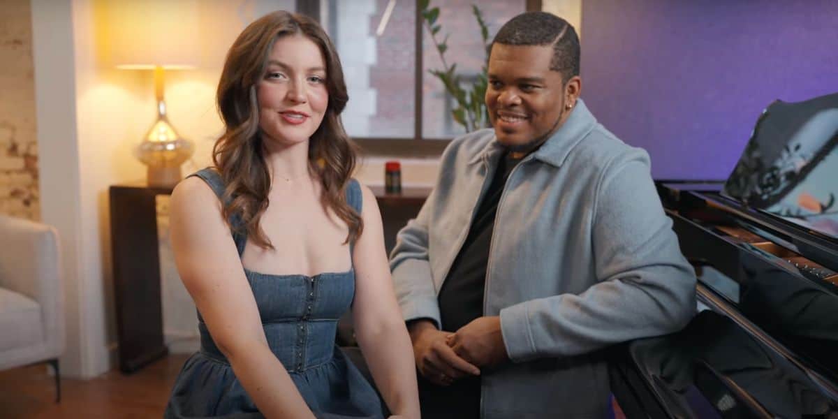 Two people sit cozily indoors near a piano, evoking a scene reminiscent of Disney's "Beauty and the Beast." The woman wears a denim dress, while the man sports a light jacket. A lamp casts a warm glow, highlighting plants in the background as they share an engaging conversation.