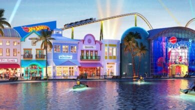 Brightly lit theme park at dusk with a waterfront. Buildings include colorful storefronts like Hot Wheels and Barbie. A roller coaster runs above. People in small boats float on the water in the foreground.