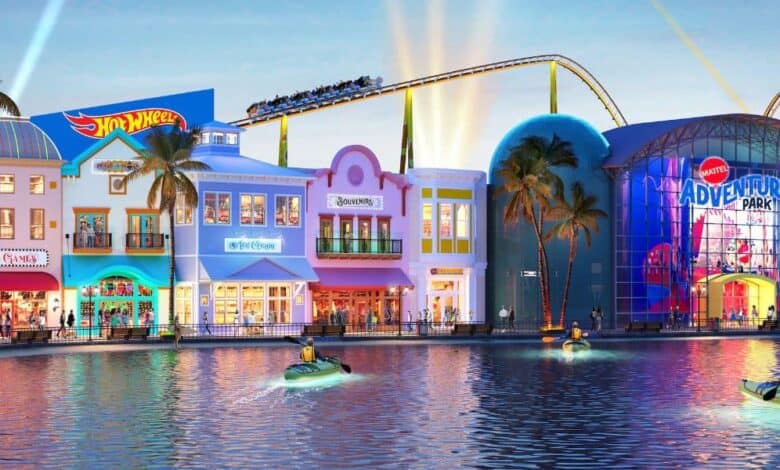 Brightly lit theme park at dusk with a waterfront. Buildings include colorful storefronts like Hot Wheels and Barbie. A roller coaster runs above. People in small boats float on the water in the foreground.