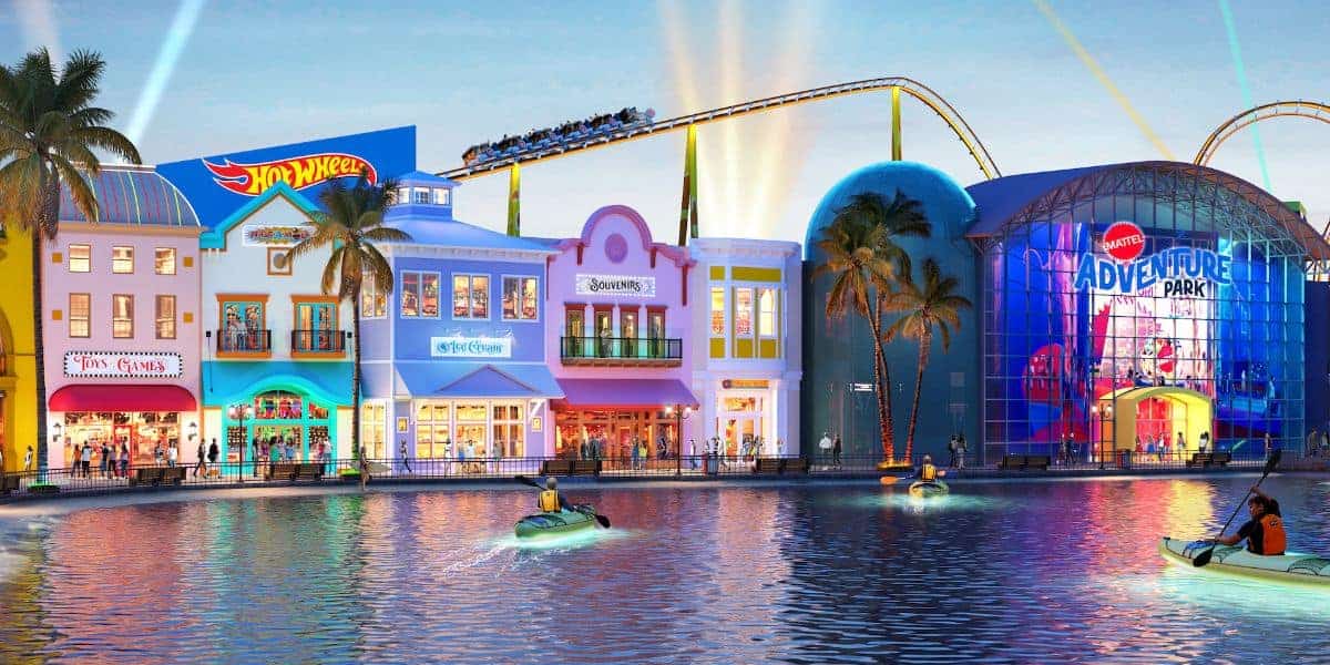 Brightly lit theme park at dusk with a waterfront. Buildings include colorful storefronts like Hot Wheels and Barbie. A roller coaster runs above. People in small boats float on the water in the foreground.