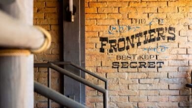 A brick wall with black text that reads, "The Frontiers Best Kept Secret." Metal railings and a wooden post are visible in the foreground.