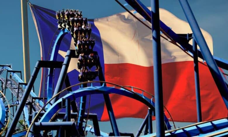 A roller coaster with people riding is in the foreground, featuring multiple loops and blue tracks. In the background, the Texas state flag waves against a clear blue sky at this Six Flags theme park.
