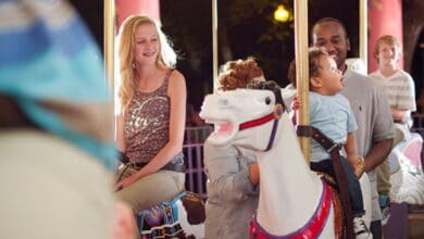 People enjoy a carousel ride at night. A smiling woman and a man holding a child sit on white carousel horses. Others can be seen in the background, surrounded by colorful decorations and soft lighting.