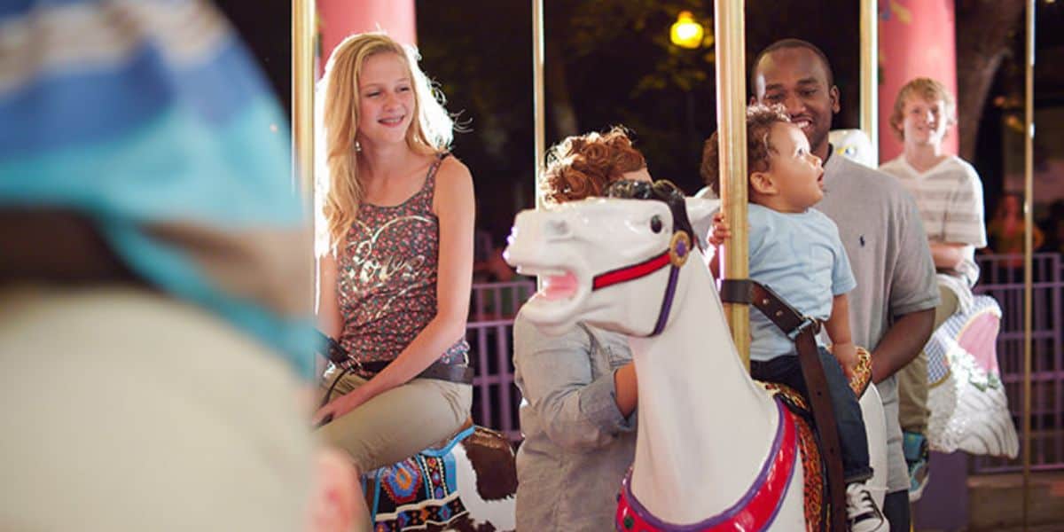 People enjoy a carousel ride at night. A smiling woman and a man holding a child sit on white carousel horses. Others can be seen in the background, surrounded by colorful decorations and soft lighting.