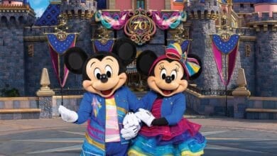 Mickey and Minnie Mouse, dressed in colorful, festive outfits, stand joyfully in front of a castle adorned with "70" banners at this Disney Park.