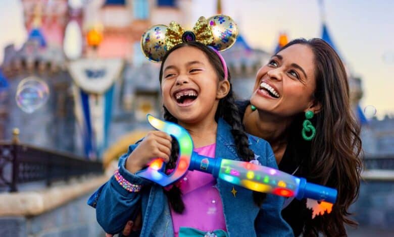 A child wearing glittery mouse ears and holding a colorful bubble wand smiles joyfully next to an adult. They stand in front of a castle adorned with banners and flags. Both appear to be enjoying the moment, with bubbles floating around them at Disneyland.