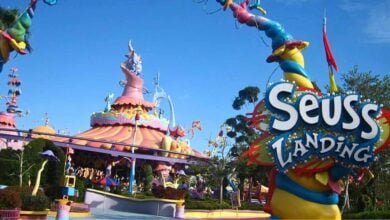 Colorful entrance to "Seuss Landing" at an amusement park, featuring whimsical architecture with twisted towers and bright colors. The sky is clear, and trees can be seen in the background.