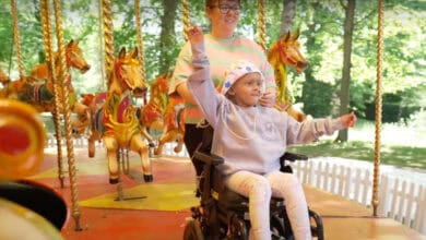 A child in a wheelchair, smiling and raising an arm, enjoys a carousel ride with a caregiver behind them. Decorative carousel horses and trees are visible in the bright background.