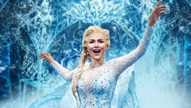 A woman dressed as Elsa from Disney’s Frozen, wearing a sparkling ice-blue gown with a long cape, stands with arms raised and smiling against a backdrop of icy patterns and snowflakes.