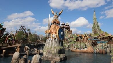 A large Viking statue holding a sword and shield stands on an island in a theme park’s epic universe, surrounded by water, Viking ships, and colorful buildings under a blue sky with scattered clouds.