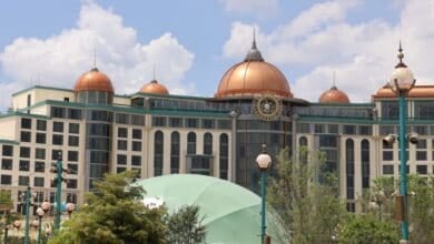 Large, elegant building with copper domes, arched windows, and a central clock stands in an epic universe, surrounded by street lamps, trees, and a green dome structure under a partly cloudy sky.
