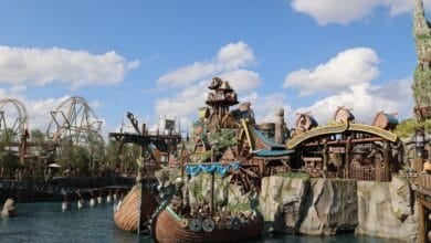 A vibrant epic universe unfolds in this theme park scene, featuring a Viking ship on water, wooden roller coasters, rocky cliffs, and whimsical buildings beneath a partly cloudy sky.